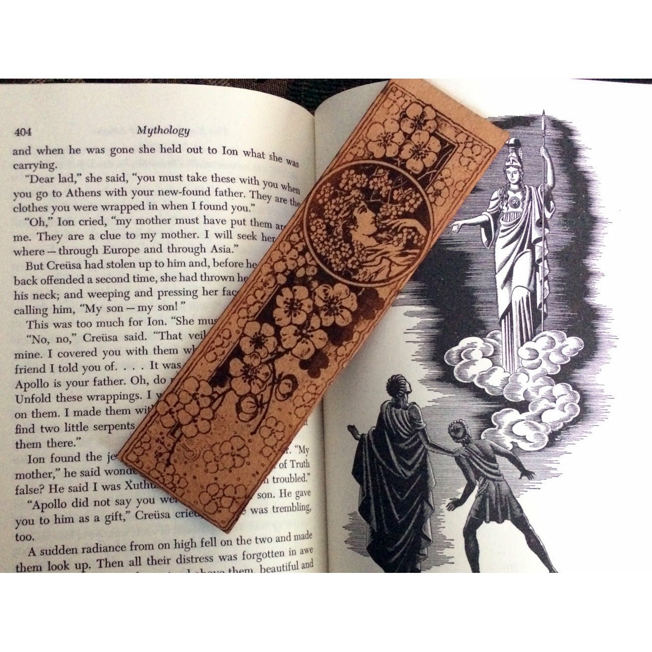 A leather bookmark of a woman with cherry blossoms that is resting on a book about mythology.