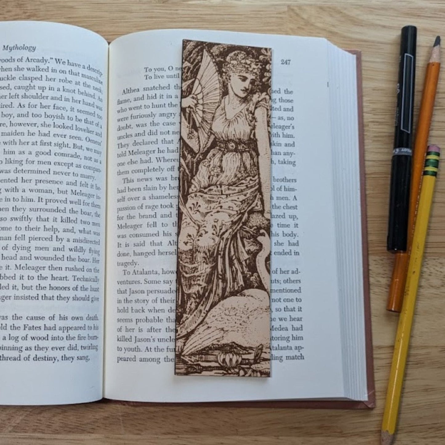 A rectangular leather bookmark with the art of a woman with a swan engraved on it. The woman is resting next to the swan while holding her fan. The bookmark is resting on the pages of an open book. Woman with a Swan Historical Leather Bookmark Bookmarks Raven King Crafts Woman with a Swan Historical Leather Bookmark Bookmarks Raven King Crafts