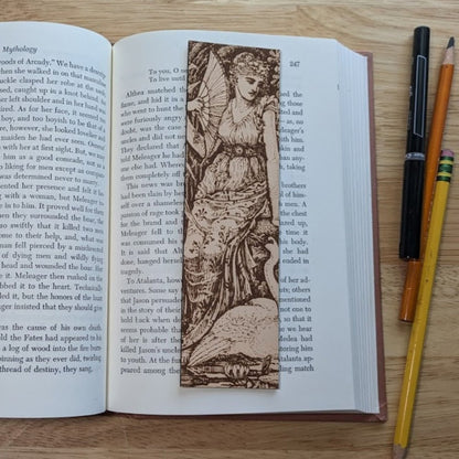 A rectangular leather bookmark with the art of a woman with a swan engraved on it. The woman is resting next to the swan while holding her fan. The bookmark is resting on the pages of an open book. Woman with a Swan Historical Leather Bookmark Bookmarks Raven King Crafts Woman with a Swan Historical Leather Bookmark Bookmarks Raven King Crafts