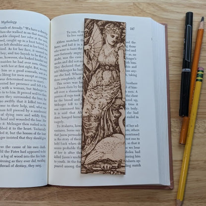 A rectangular leather bookmark with the art of a woman with a swan engraved on it. The woman is resting next to the swan while holding her fan. The bookmark is resting on the pages of an open book.