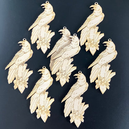 A photograph of 8 wooden raven ornament blanks sitting on a black background. The ravens are sitting on crystal clusters.